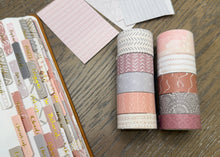 Load image into Gallery viewer, Two rolls of decorative washi tape with pink, gray, and white patterns, displayed on a wooden surface next to a notebook filled with labeled tabs and patterned paper swatches.
